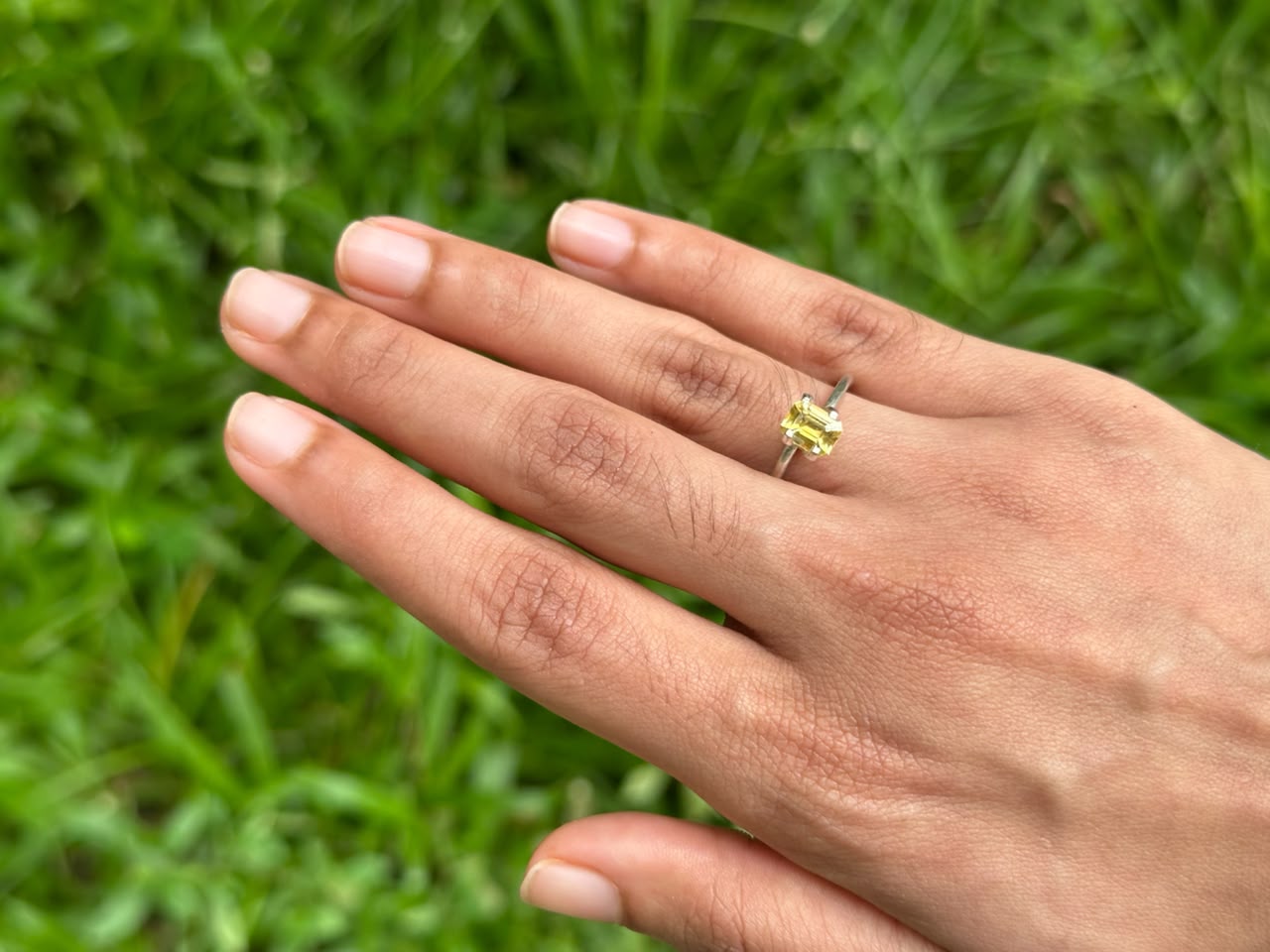 Hand wearing a ring featuring a 1.17 ct Emerald-Cut Ceylon Yellow Sapphire on green grass background.