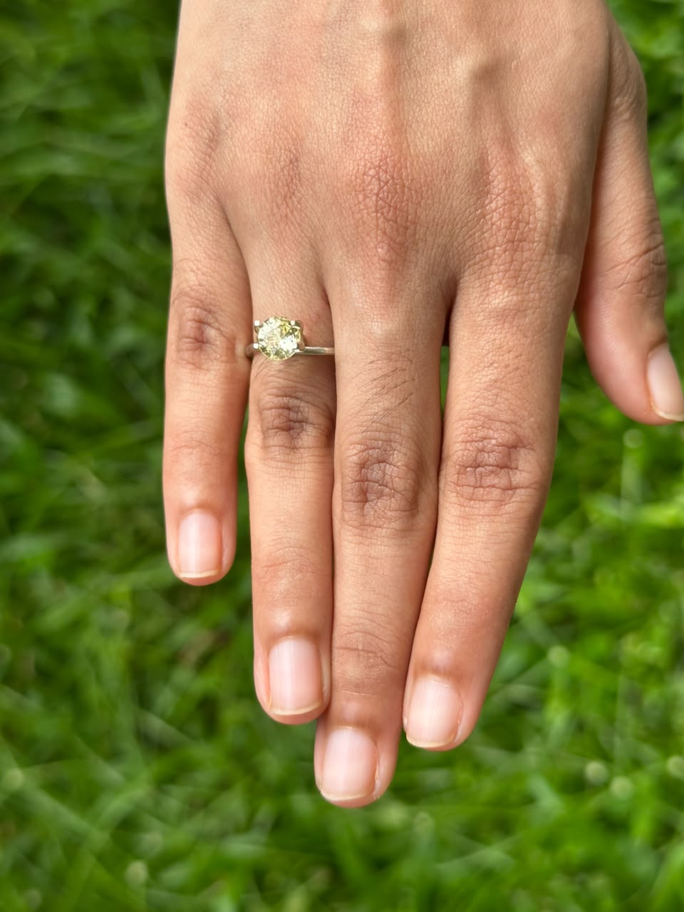 Hand wearing a 1.44 ct Round Ceylon Yellow Sapphire engagement ring on green grass.
